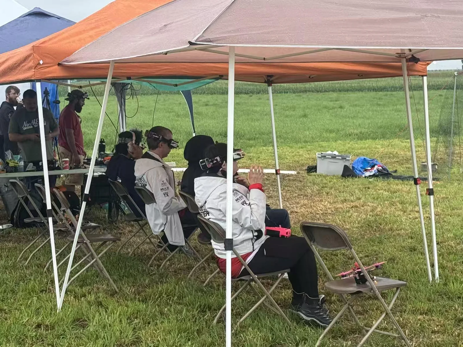 FPV drone racing pilots seated beneath event tents beside a race field